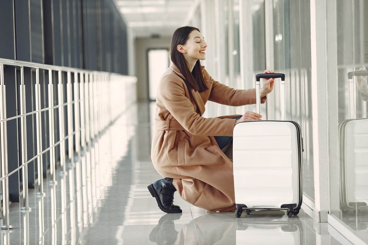 Side view full length of cheerful young female passenger in stylish beige coat with suitcase hunkering down and looking out glass wall in contemporary airport corridor