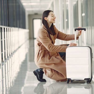 Side view full length of cheerful young female passenger in stylish beige coat with suitcase hunkering down and looking out glass wall in contemporary airport corridor
